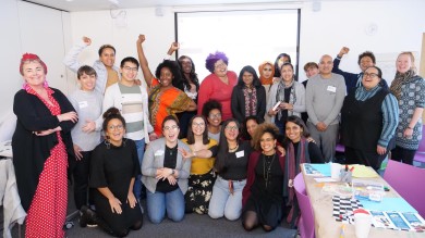 A group photo of 24 workshop attendees, mostly women of colour, all skin tones and hair colours with colourful outfits, big smiles and positive energy. An Asian woman has an orange hijab, while a black woman is wearing a dashiki. few of them are holding up their right fists or a peace sign.
